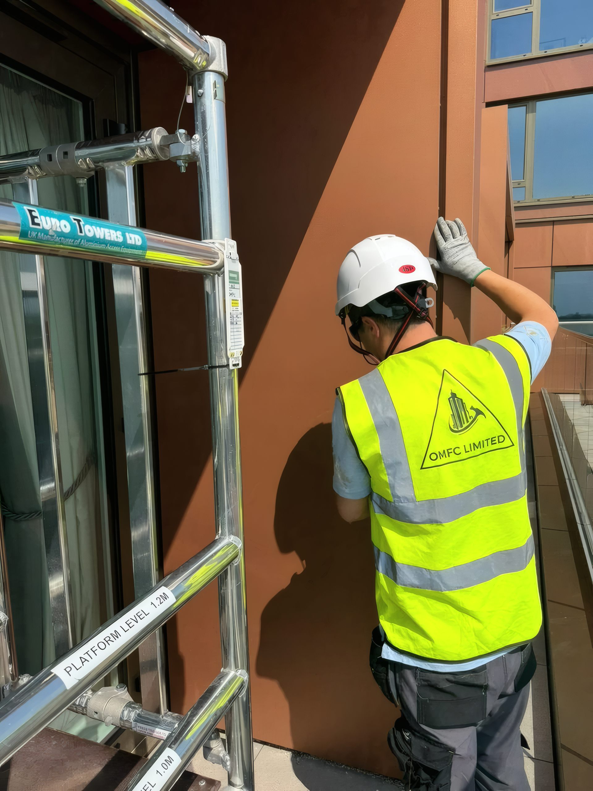 An installer in an OMFC Limited hi-vis vest fitting terracotta-finish cladding panels from an access tower.